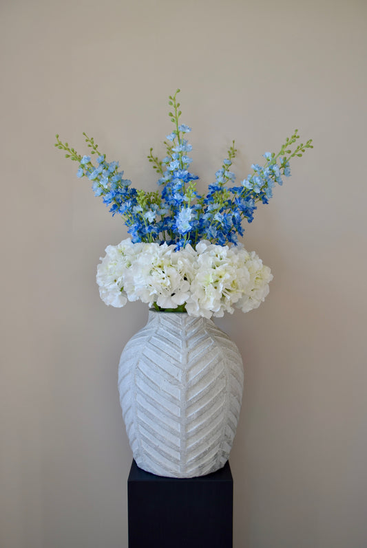 White artificial hydrangea and blue delphinium flower arrangement placed in a chevron style vase on a black pedestal against a plain wall. 