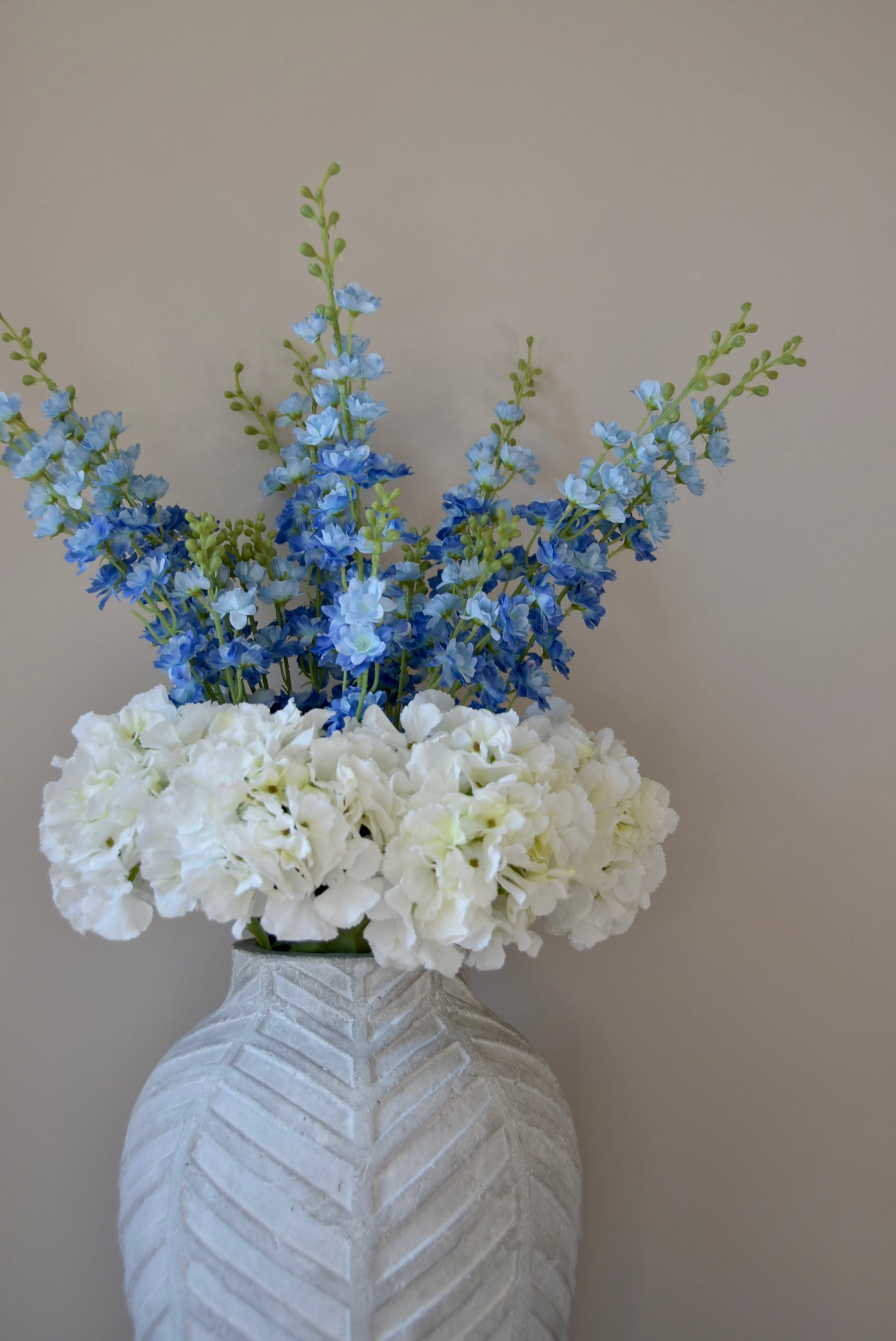 White artificial hydrangea and blue delphinium flower arrangement placed in a chevron style vase against a plain wall. 
