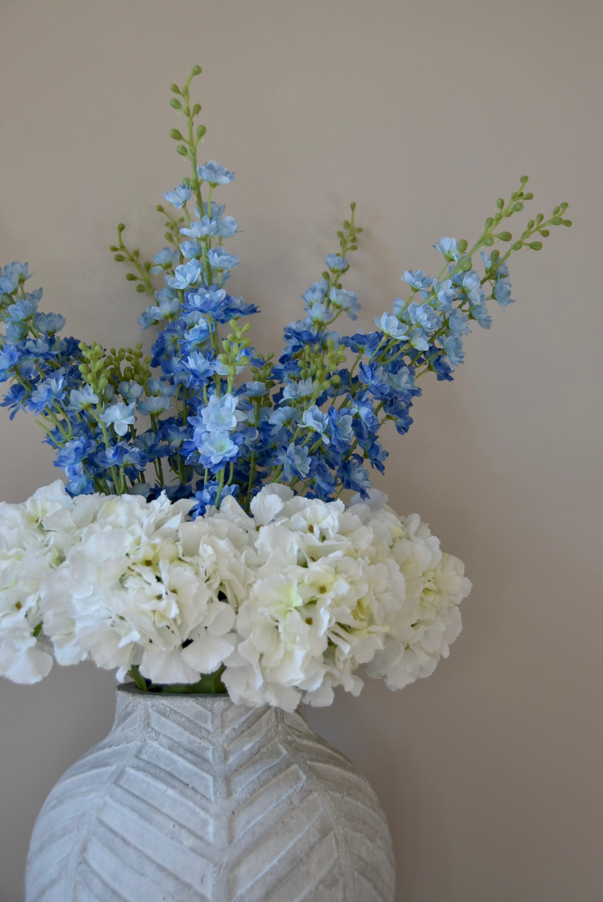 White artificial hydrangea and blue delphinium flower arrangement placed in a chevron style vase against a plain wall. 