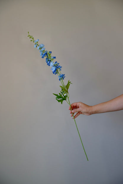 A hand holding a single artificial blue delphinium stem.