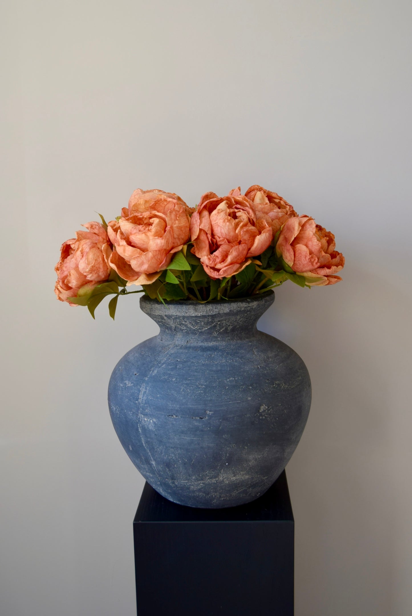 Grey textured vase with orange peonies on a black stand against a plain background