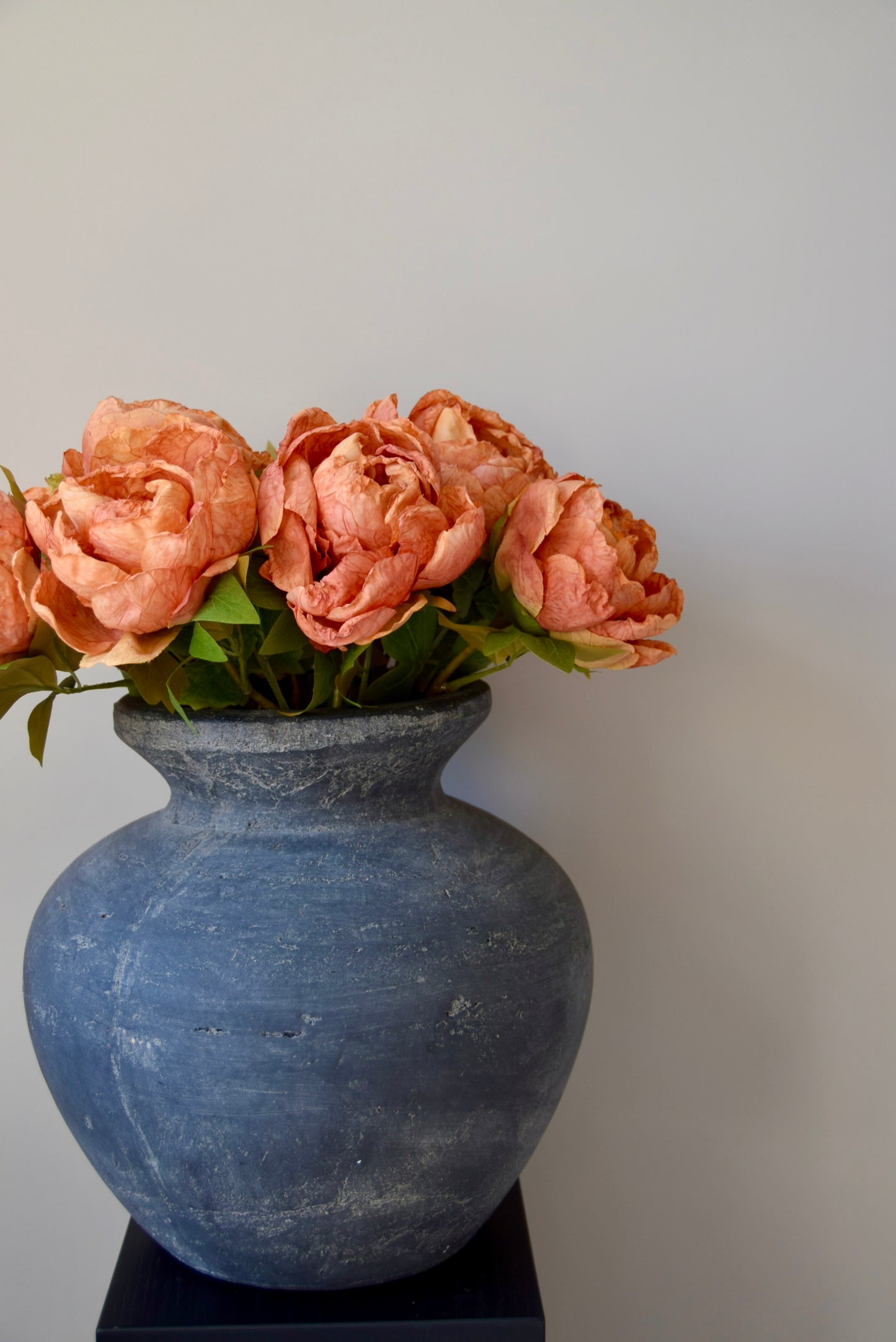 Grey textured vase with orange peonies on a black stand against a plain background