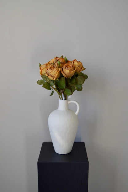 White vase with honey roses on a black pedestal against a plain wall