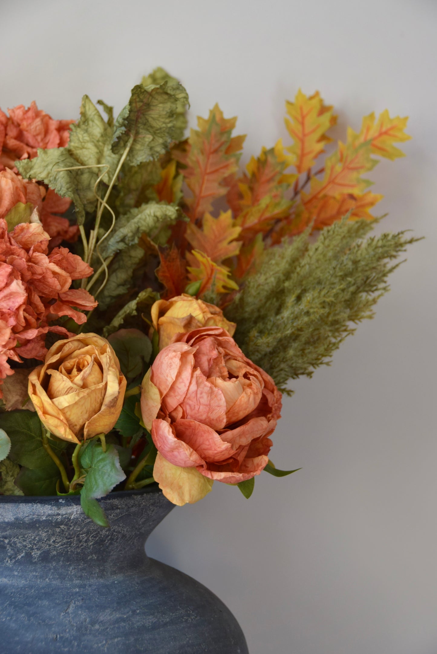 Autumn-themed floral arrangement with pink, orange, and green leaves in a dark vase against a light grey background.