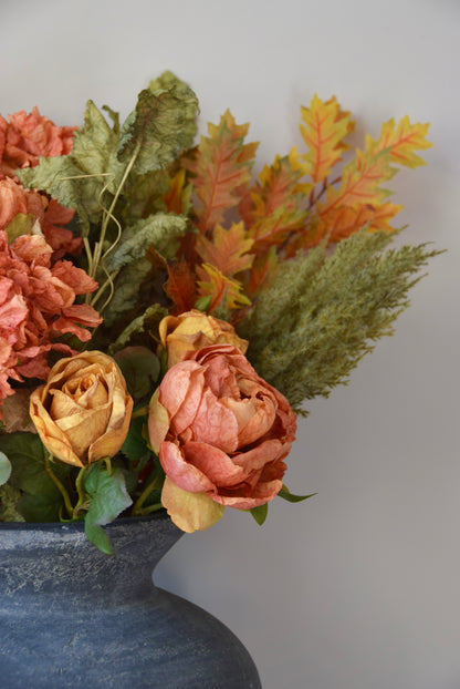 Autumn-themed floral arrangement with pink, orange, and green leaves in a dark vase against a light grey background.