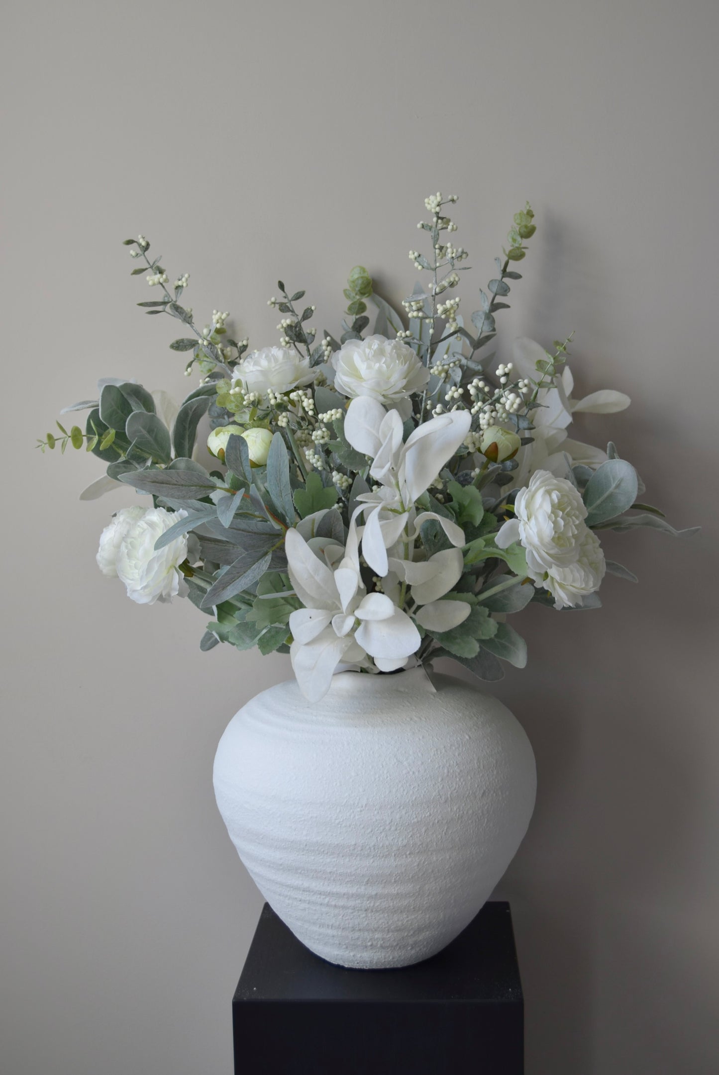 White vase with a bouquet of frosted winter flowers on a black pedestal against a grey wall