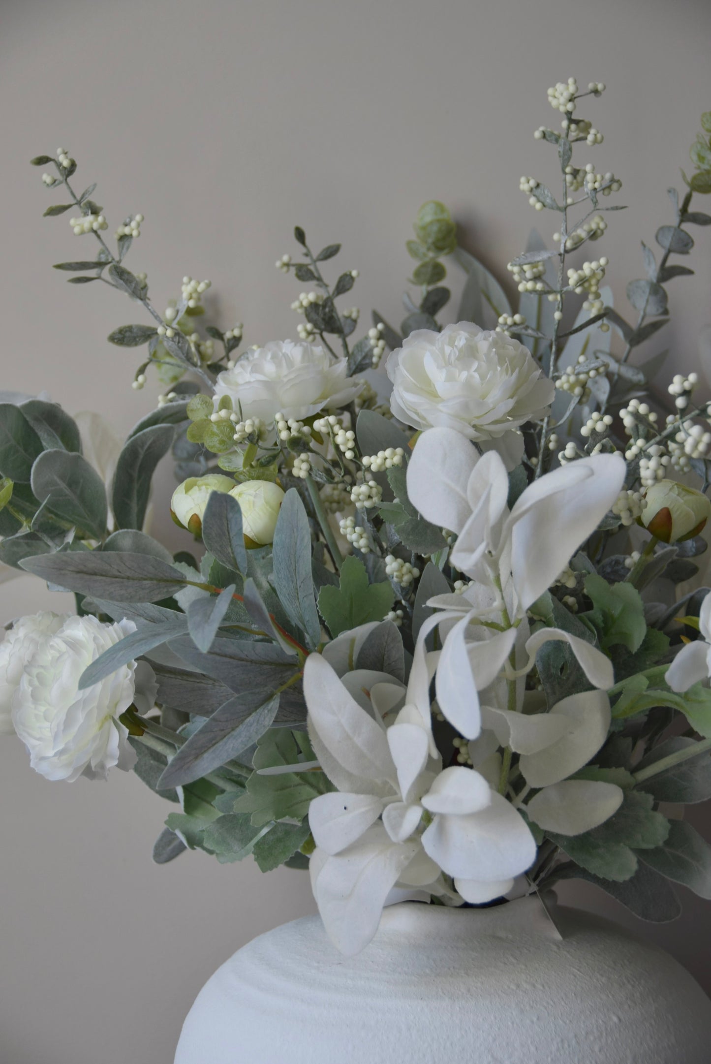 Faux floral arrangement with white flowers and greenery in a white vase against a neutral background