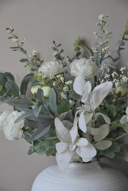 Faux floral arrangement with white flowers and greenery in a white vase against a neutral background