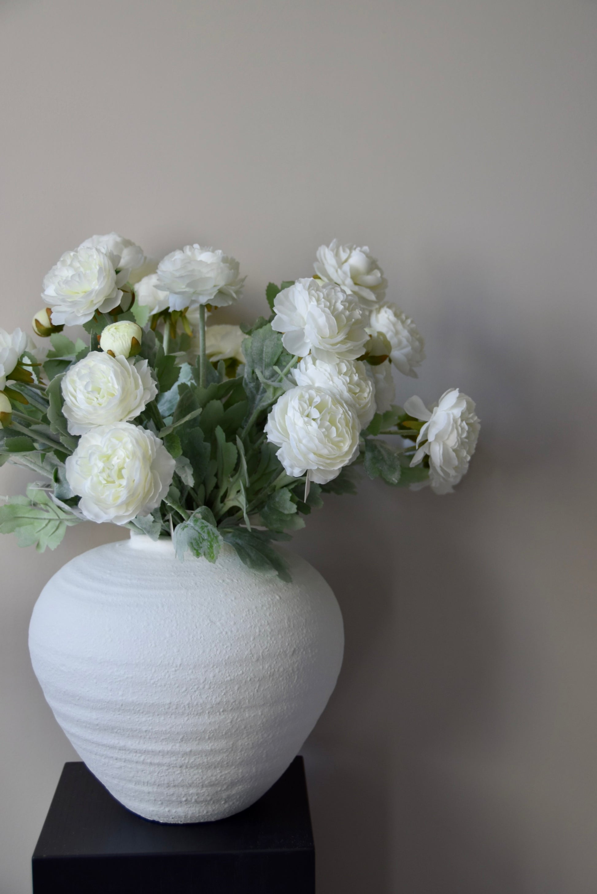White vase with white flowers on a black stand against a plain wall