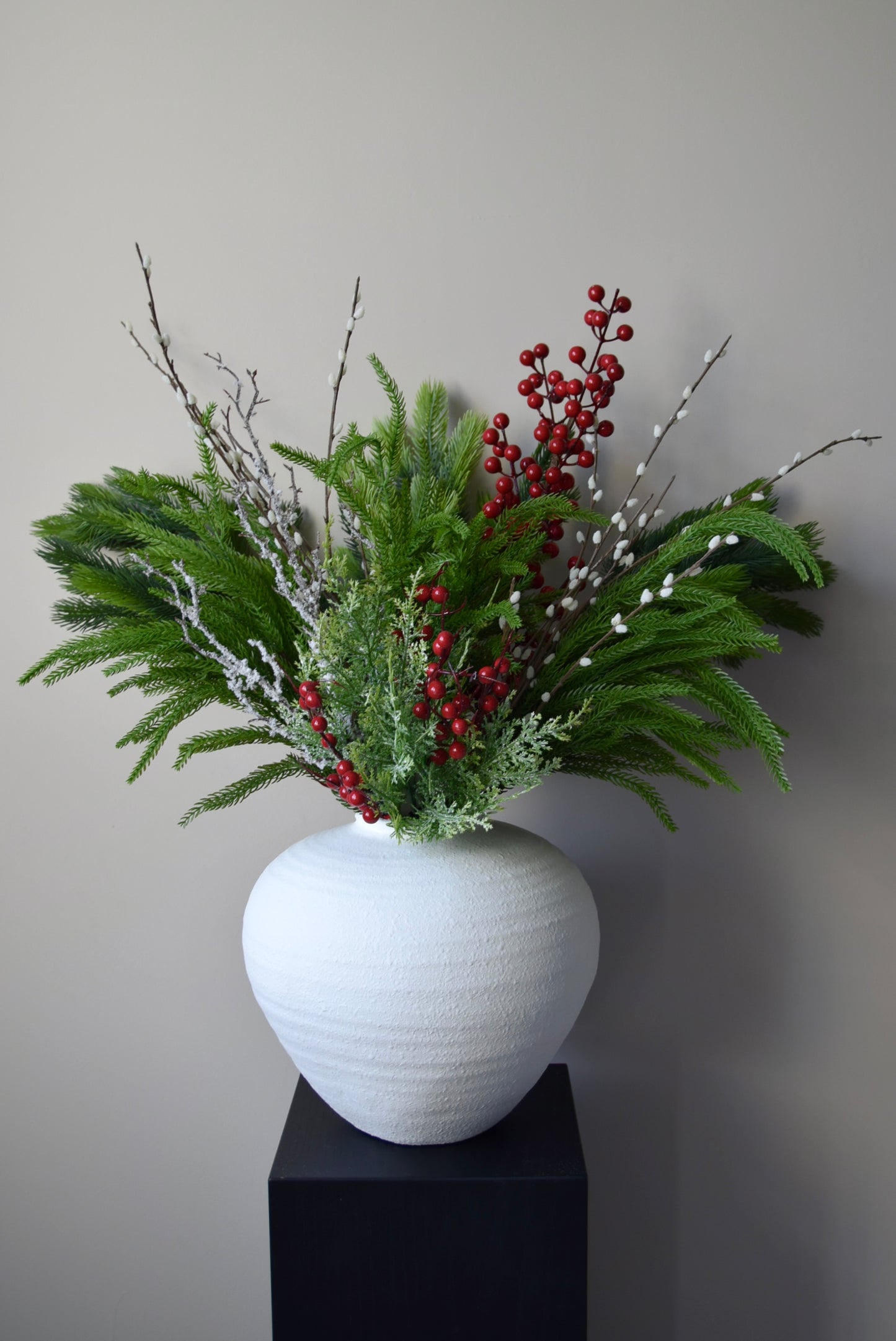 White vase with a festive green and red floral arrangement on a black stand against a plain background