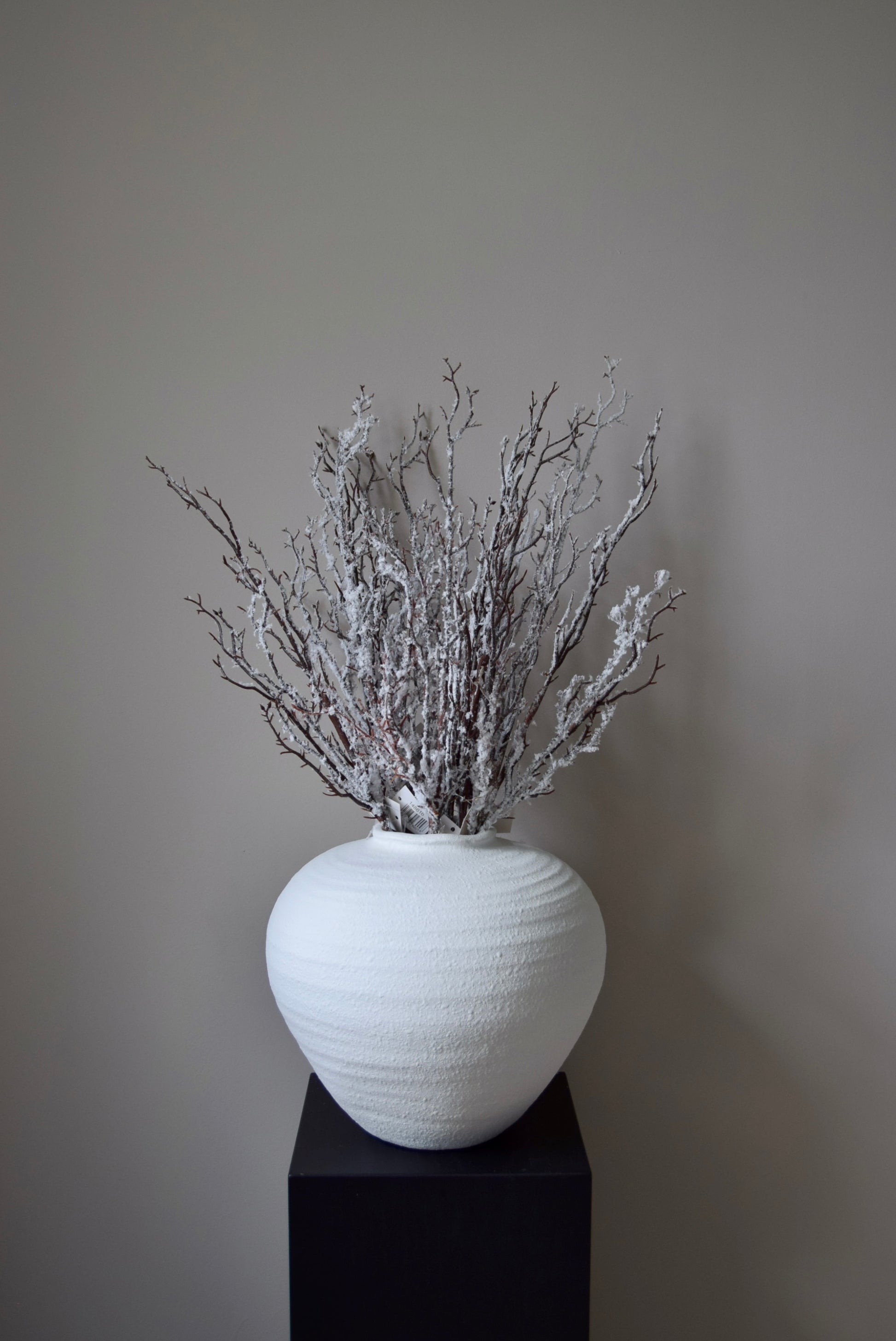 White textured vase with dried snowy branches on a black stand against a plain wall