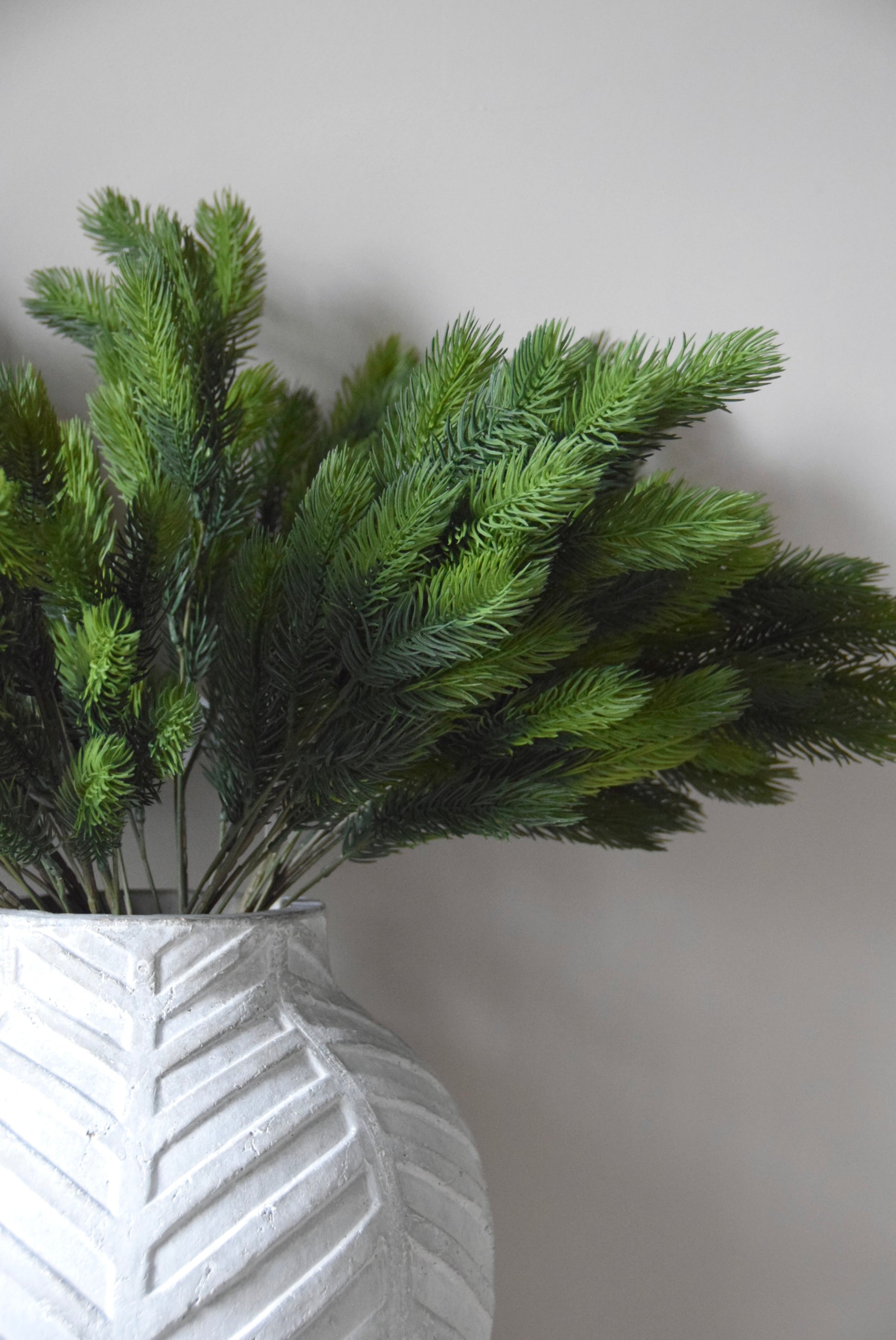 Green artificial fir branch in a textured white vase against a plain background