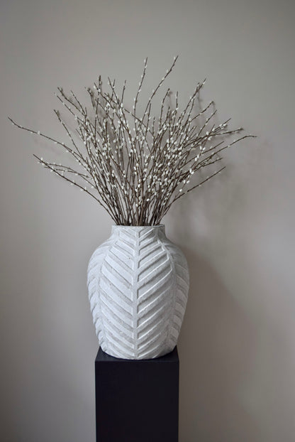 White textured vase with dried branches on a black stand against a plain background