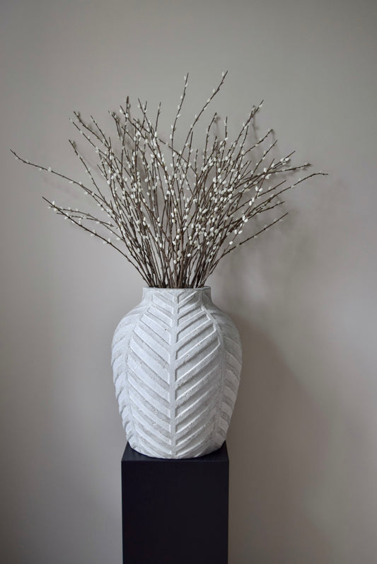 White textured vase with dried branches on a black stand against a plain background