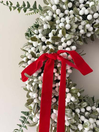 Decorative wreath with greenery, white berries, and a red ribbon on a neutral background