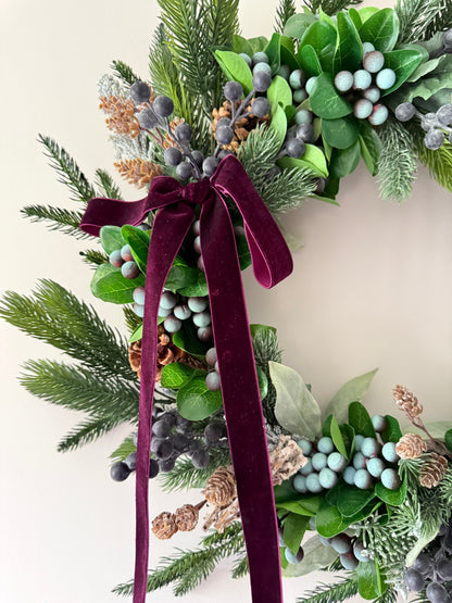 Decorative wreath with greenery, berries, and a burgundy ribbon on a light background