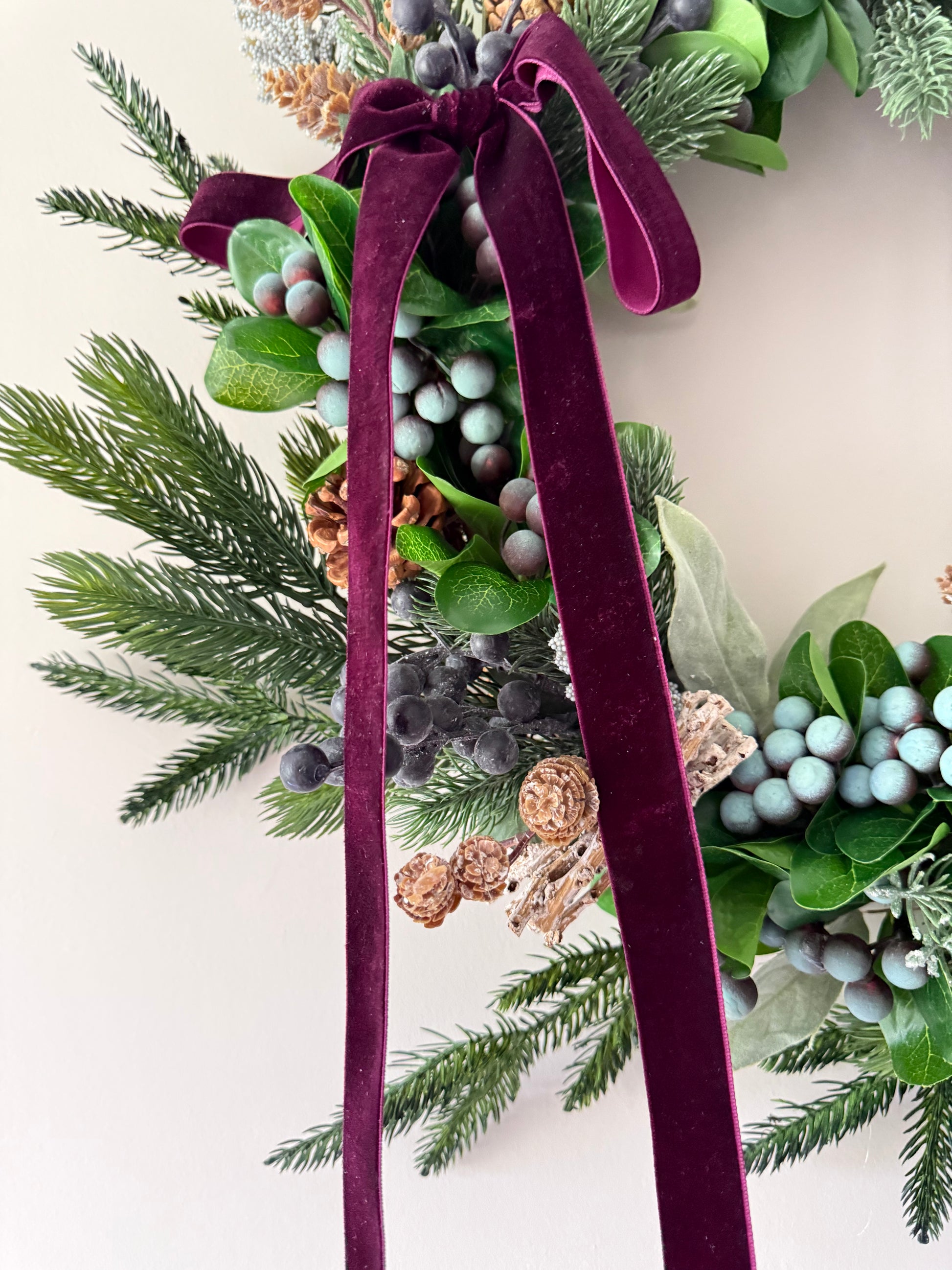 Decorative wreath with greenery, berries, and a burgundy ribbon on a light background