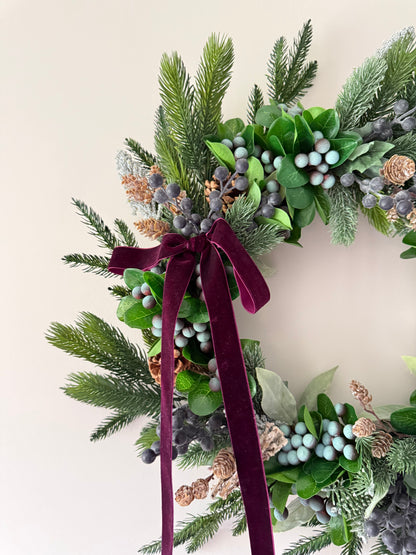 Decorative wreath with greenery, berries, and a purple ribbon on a light background