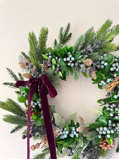 Decorative wreath with greenery, berries, and a burgundy ribbon on a light background