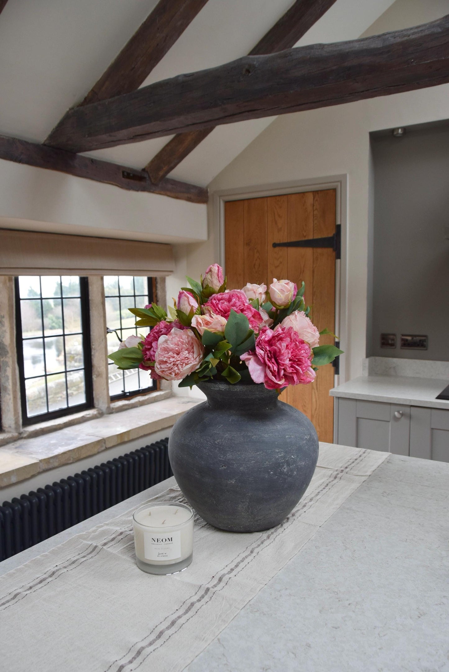 Grey antique vase placed on kitchen counter top with an arrangement of pale pink and hot pink peonies. Arranged in a country style kitchen for home decor.