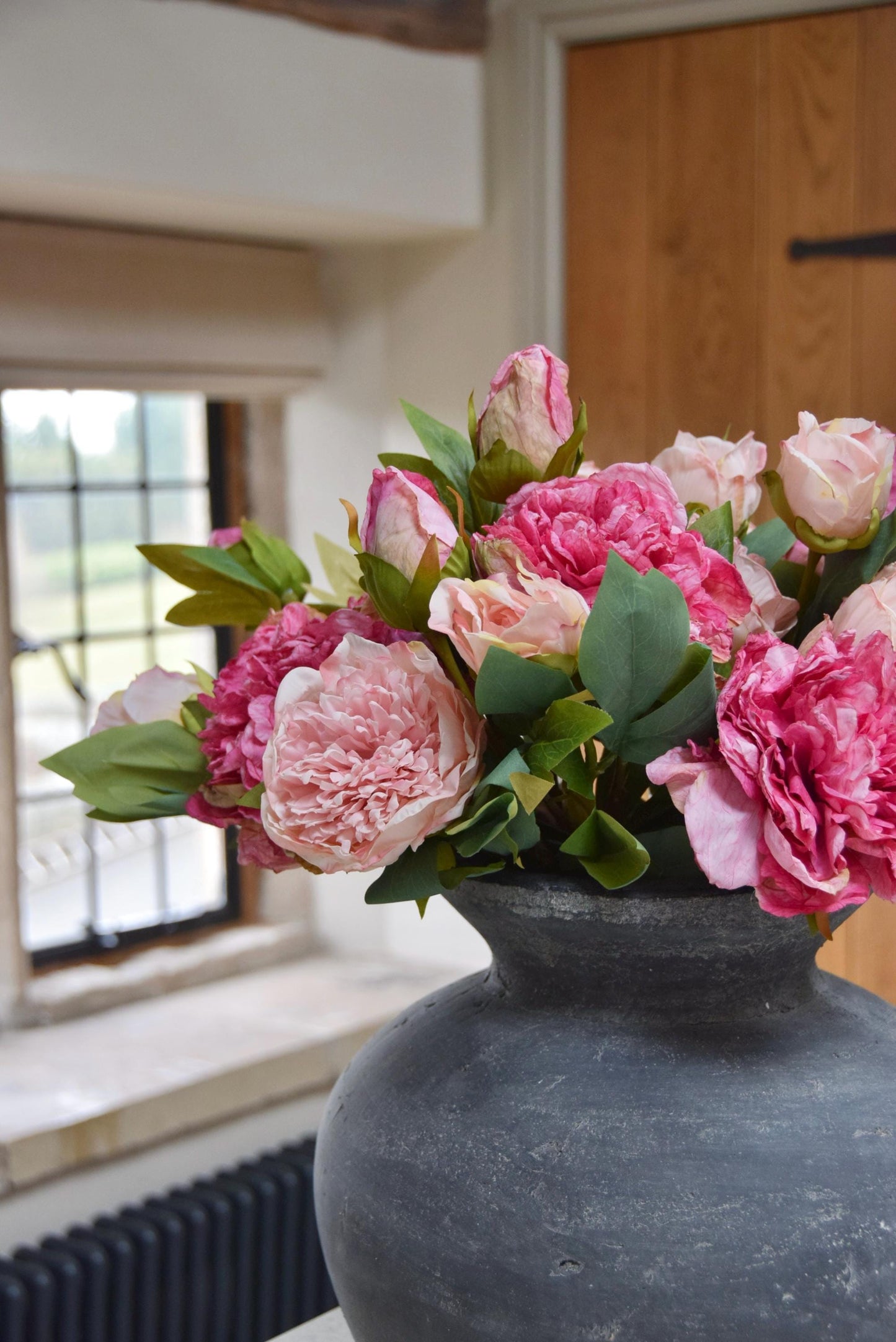 Grey antique vase placed on kitchen counter top with an arrangement of pale pink and hot pink peonies. Arranged in a country style kitchen for home decor.