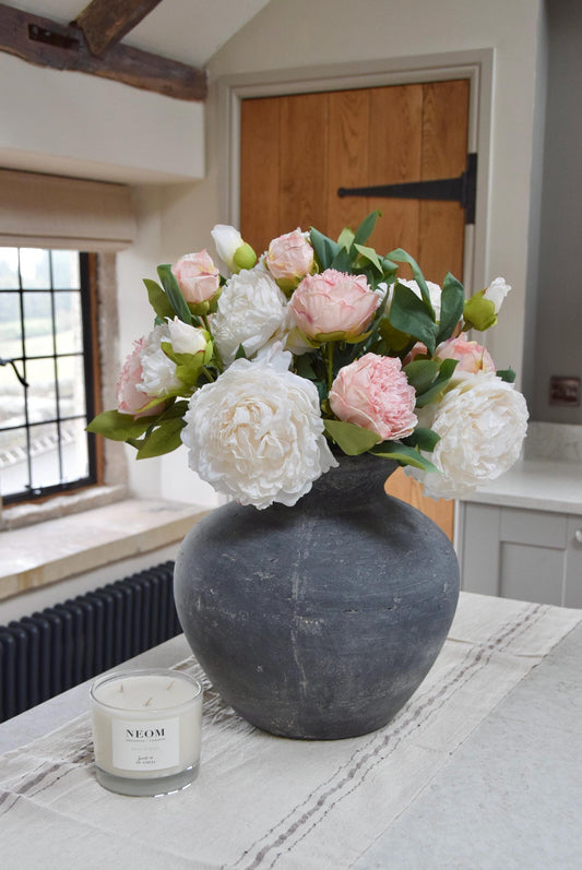 Grey antique vase placed on a kitchen counter top with an arrangement of pink and cream peonies. Arranged in a country style kitchen for home decor.
