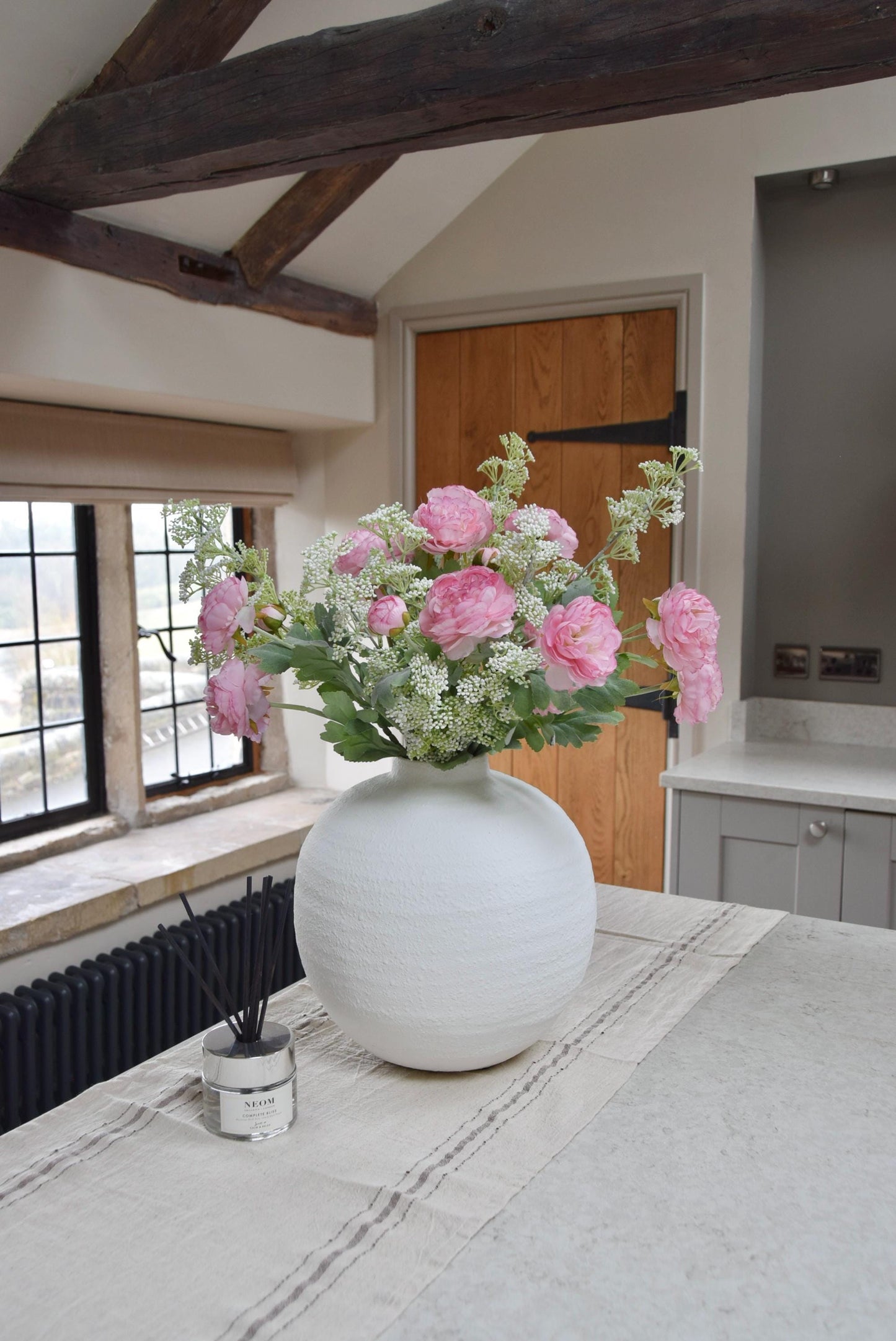 White sphere vase placed on a kitchen countertop with an arrangement of pale pink ranunculuses and white mini bud stems. Displayed in a country style kitchen for home decor.