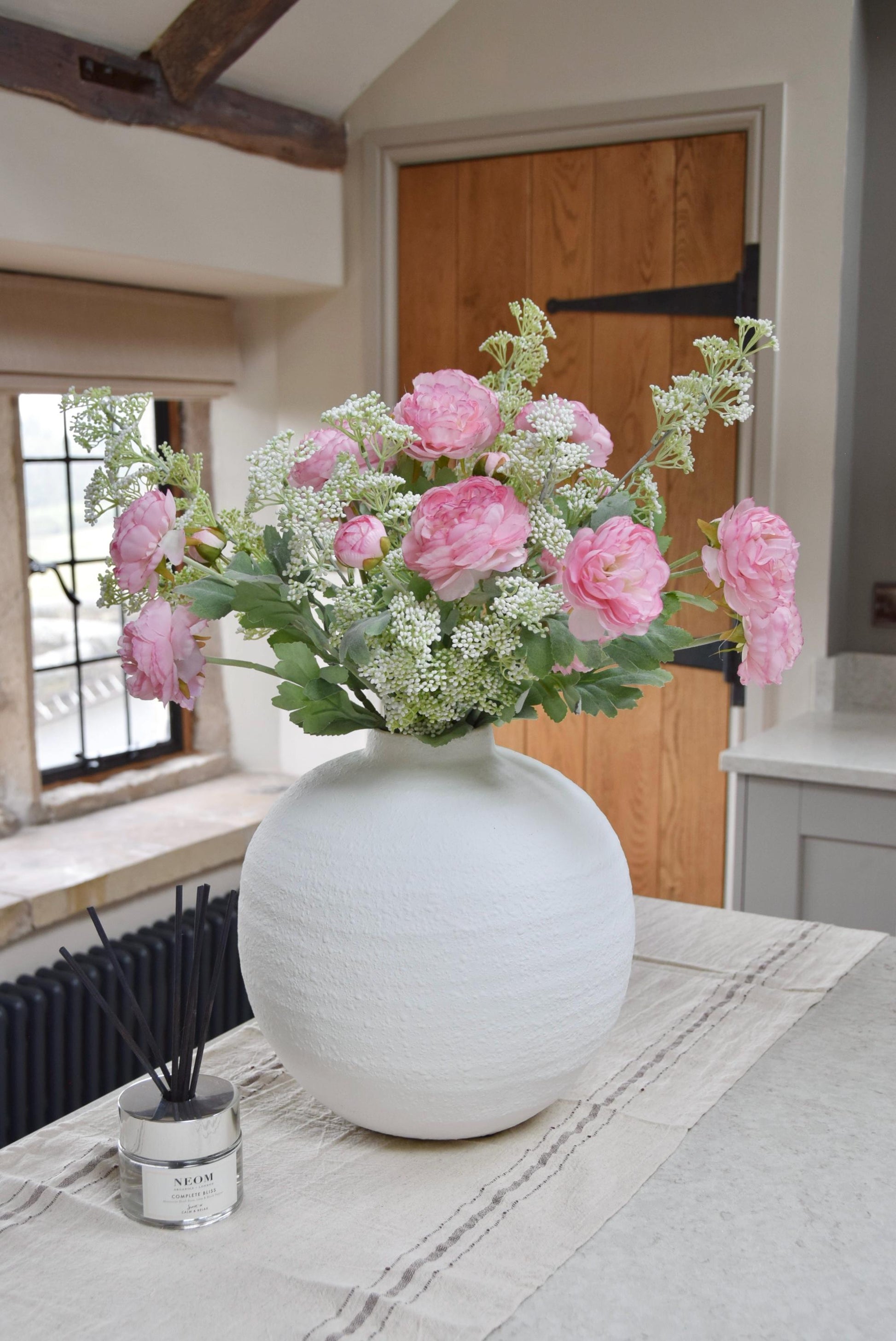 White sphere vase placed on a kitchen countertop with an arrangement of pale pink ranunculuses and white mini bud stems. Displayed in a country style kitchen for home decor.