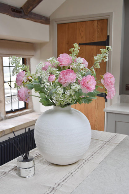 White sphere vase placed on a kitchen countertop with an arrangement of pale pink ranunculuses and white mini bud stems. Displayed in a country style kitchen for home decor.