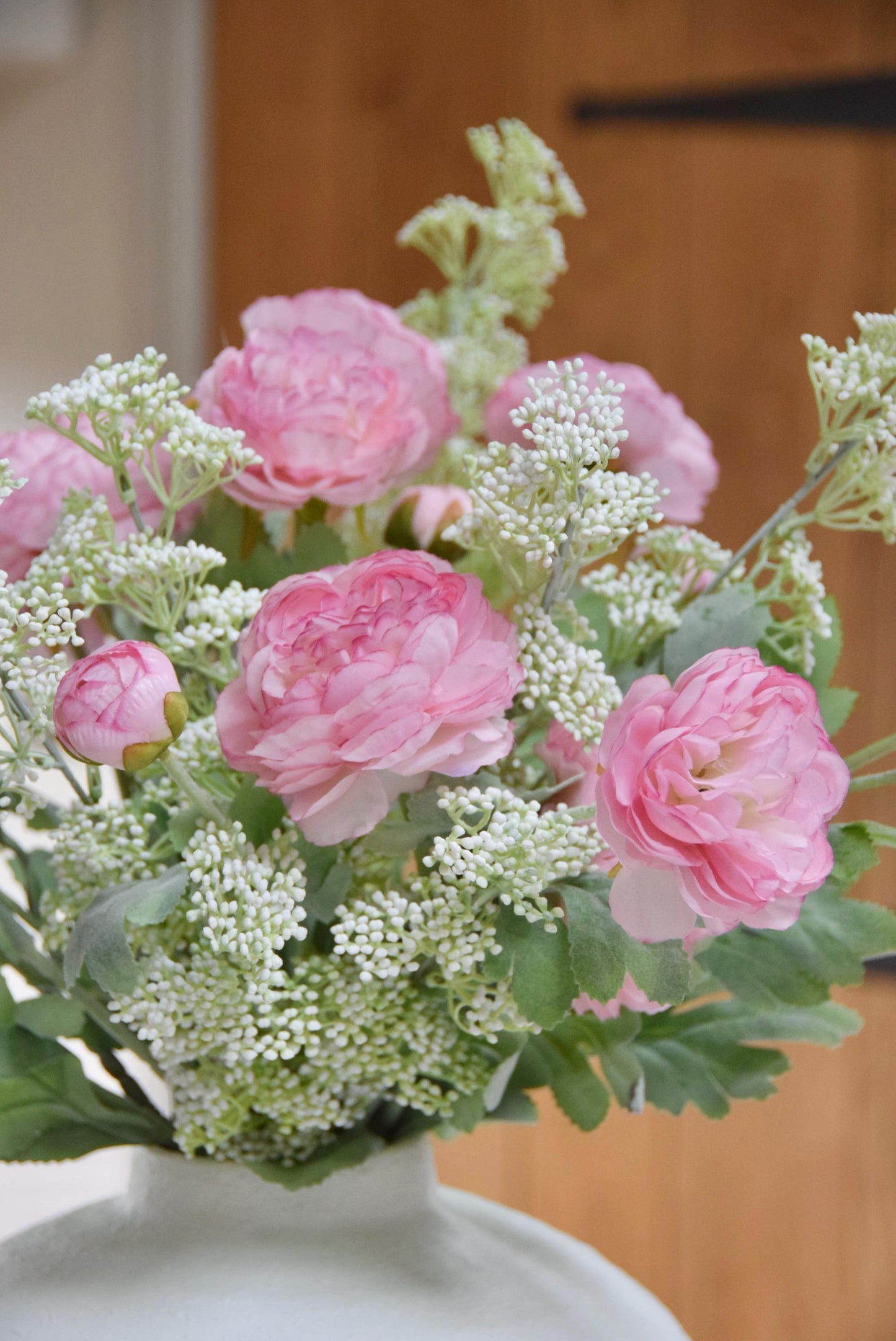 White sphere vase placed on a kitchen countertop with an arrangement of pale pink ranunculuses and white mini bud stems. Displayed in a country style kitchen for home decor.