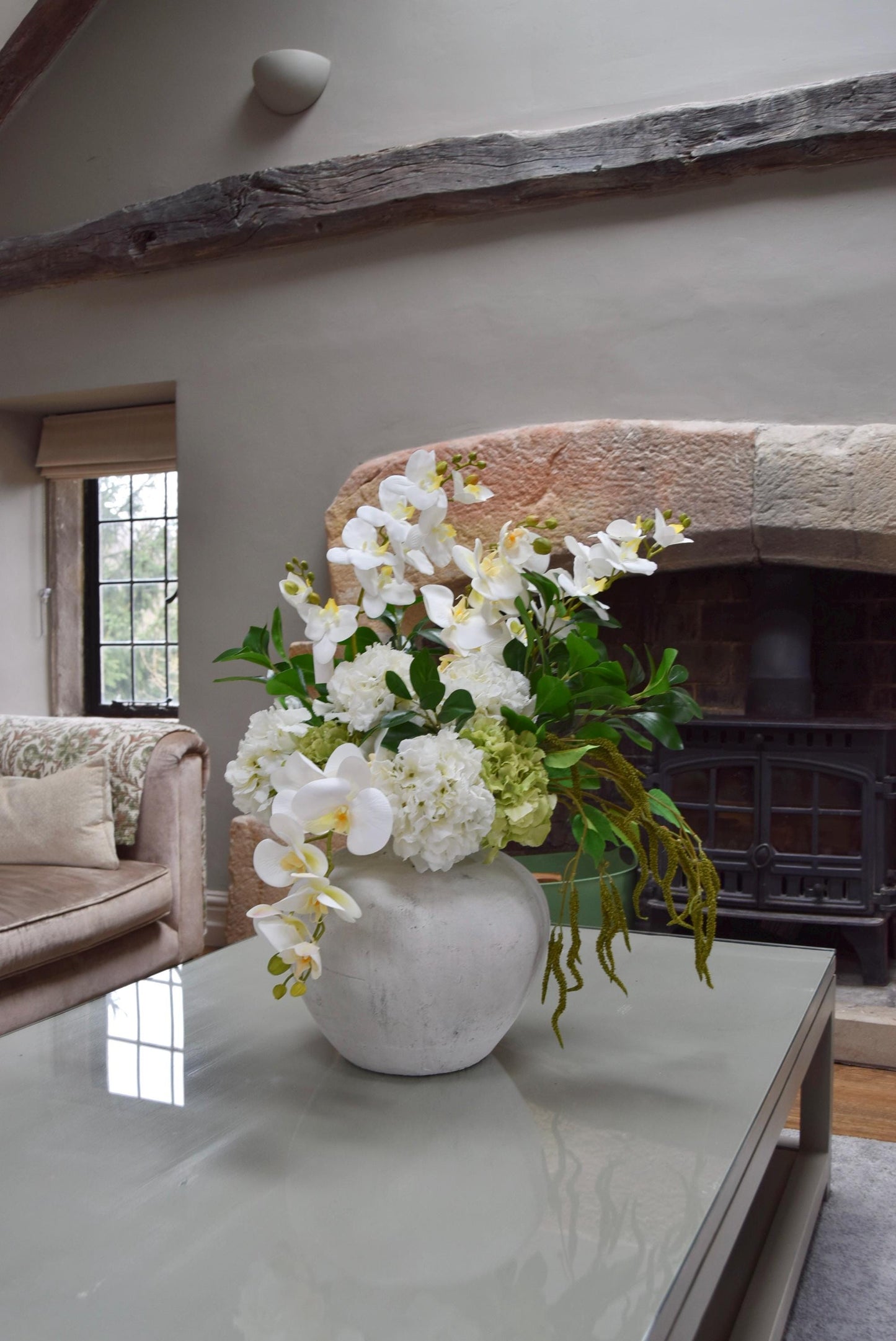 Stone antique vase placed on a kitchen countertop with an arrangement of white orchids, white hydrangeas, green hydrangeas and green foliage. Displayed in a country style kitchen for home decor.
