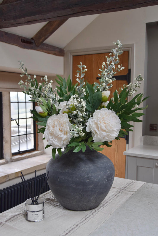 Grey antique vase placed on a kitchen countertop with artificial ruscus foliage and faux cream peony flowers. Arranged in a country style kitchen for home decor.
