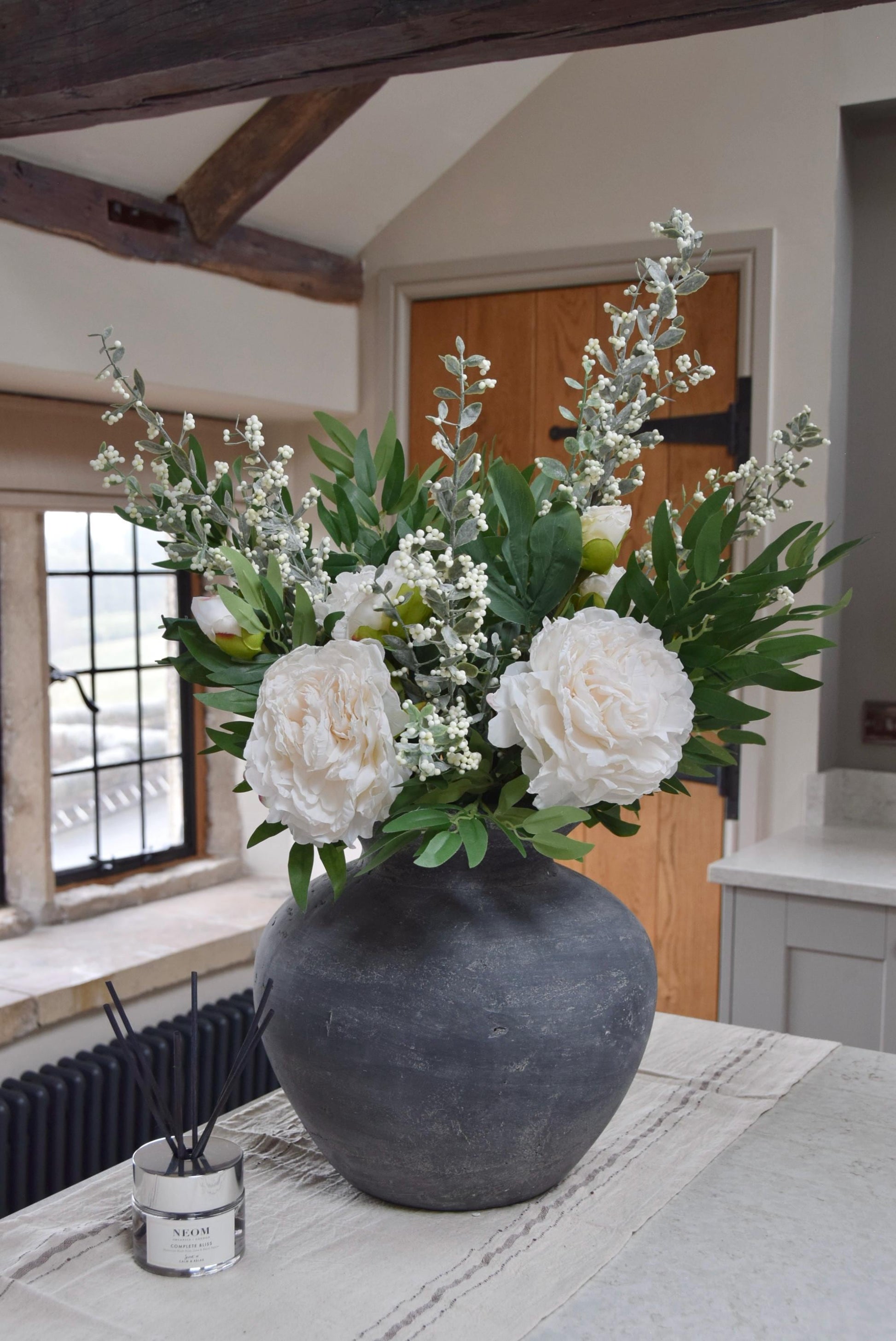 Grey antique vase placed on a kitchen countertop with artificial ruscus foliage and faux cream peony flowers. Arranged in a country style kitchen for home decor.