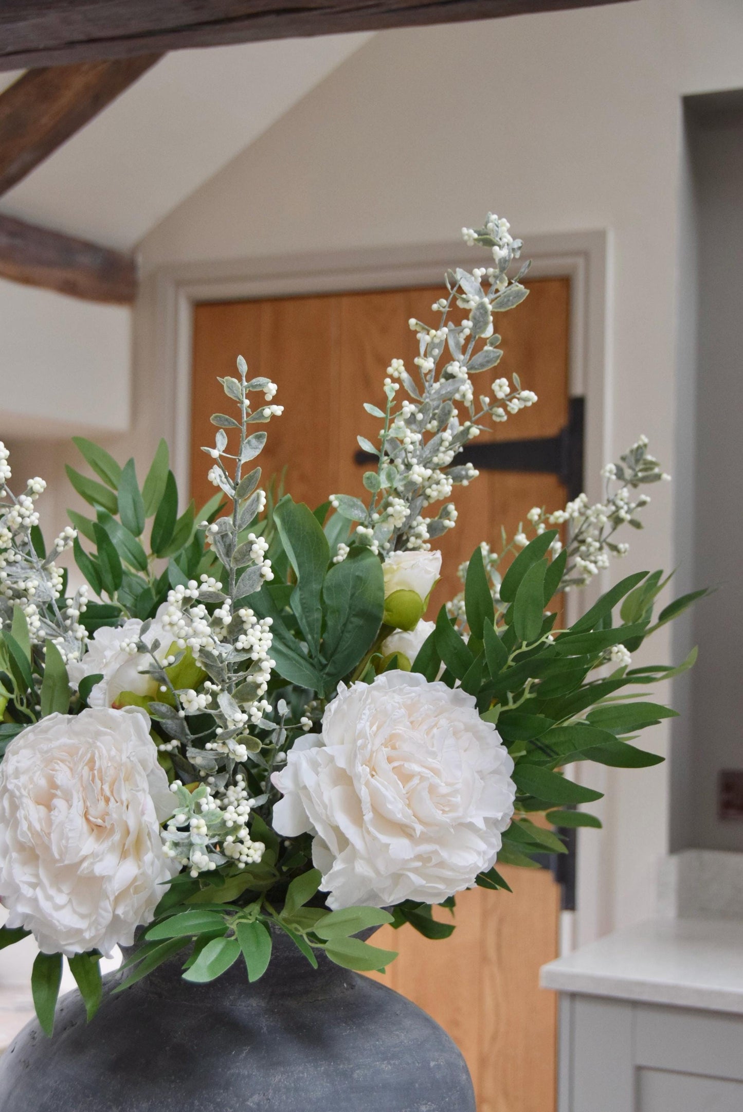 Grey antique vase placed on a kitchen countertop with artificial ruscus foliage and faux cream peony flowers. Arranged in a country style kitchen for home decor.