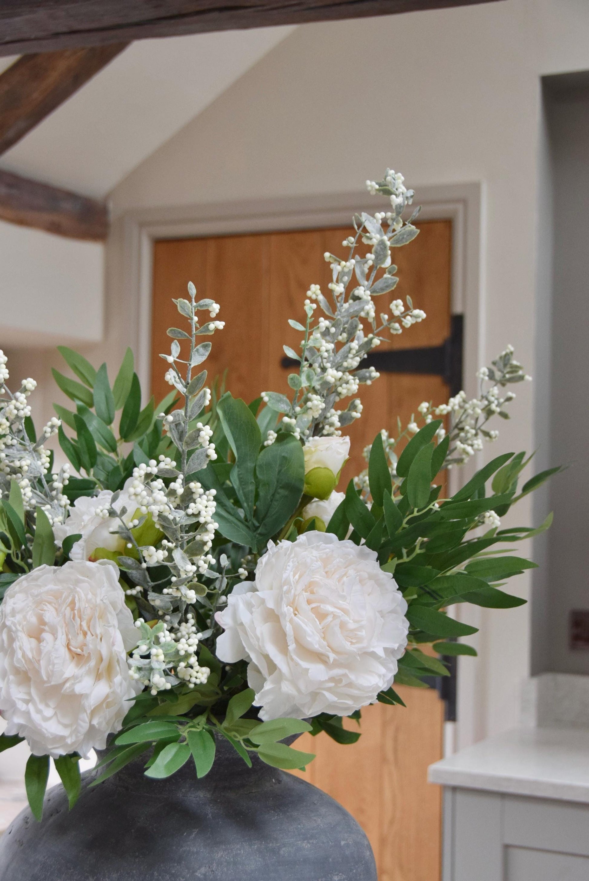 Grey antique vase placed on a kitchen countertop with artificial ruscus foliage and faux cream peony flowers. Arranged in a country style kitchen for home decor.
