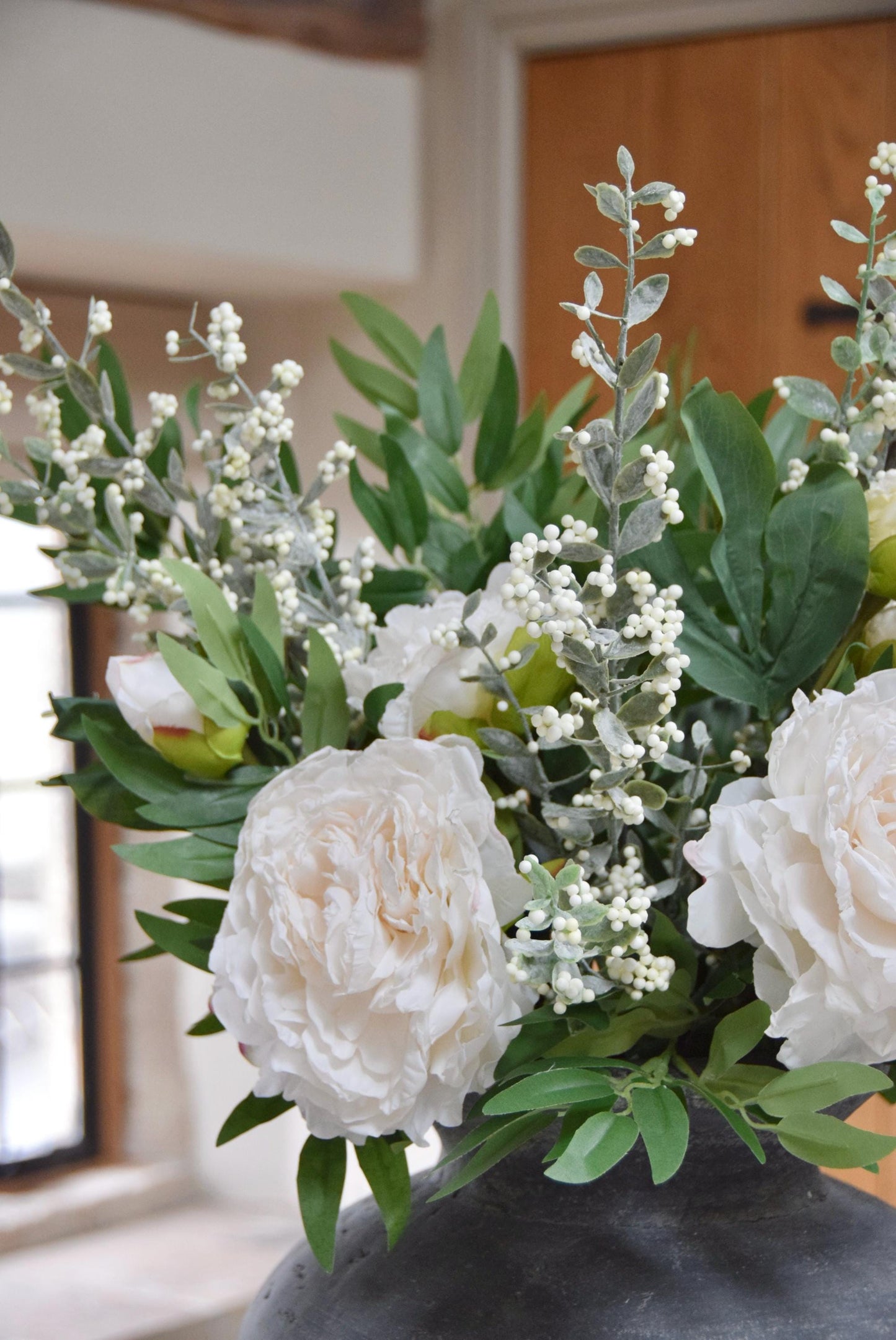 Grey antique vase placed on a kitchen countertop with artificial ruscus foliage and faux cream peony flowers. Arranged in a country style kitchen for home decor.