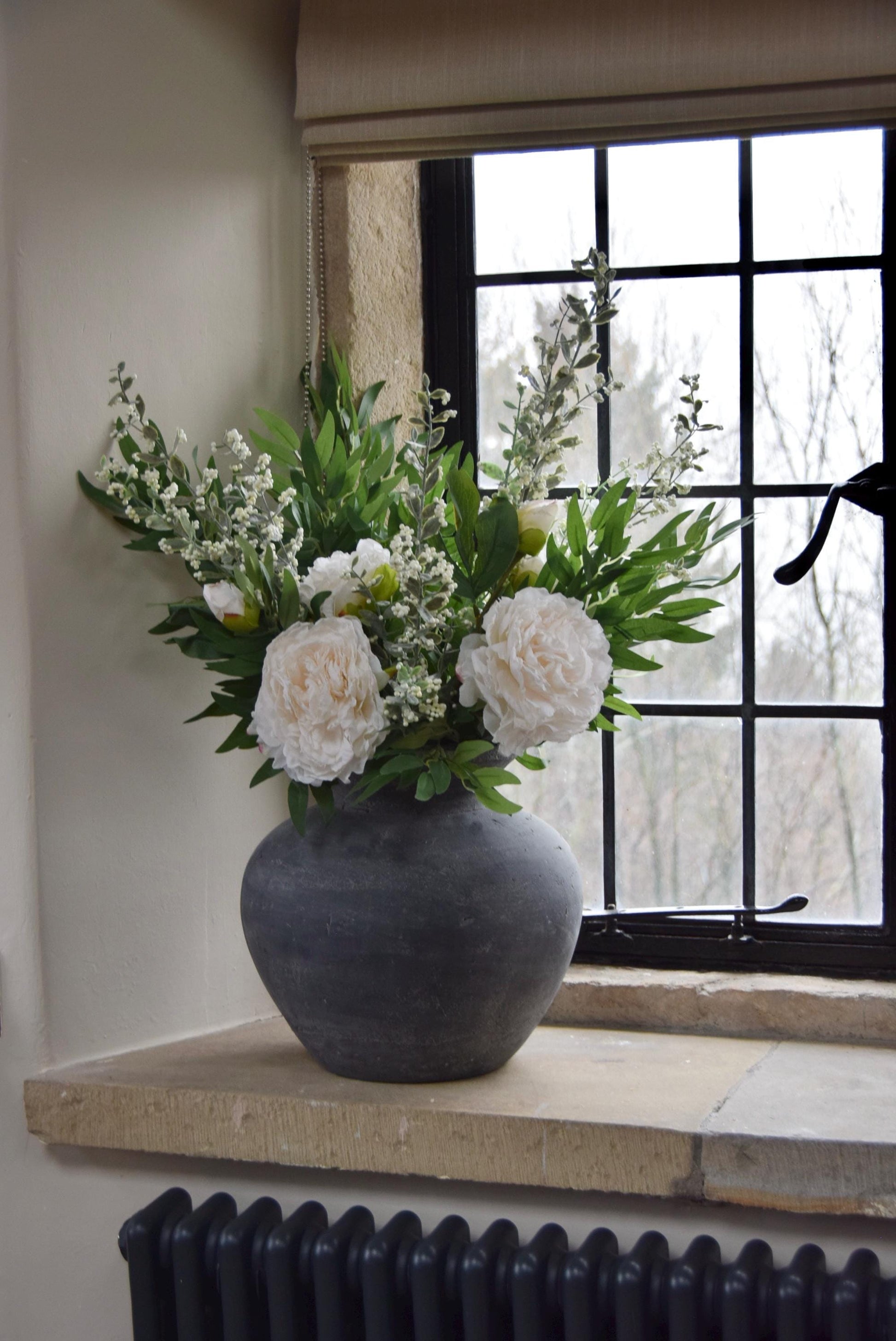 Grey antique vase placed on a stone windowsill with artificial ruscus foliage and faux cream peony flowers. Arranged in a country style kitchen for home decor.