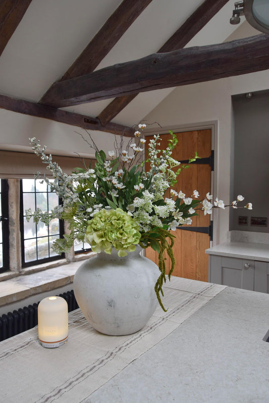 Stone antique vase placed on a kitchen countertop with an arrangement of green and white mixed flowers with lots of foliage. Displayed in a country style kitchen for home decor.Stone antique vase placed on a kitchen countertop with an arrangement of green and white mixed flowers with lots of foliage. Displayed in a country style kitchen for home decor.