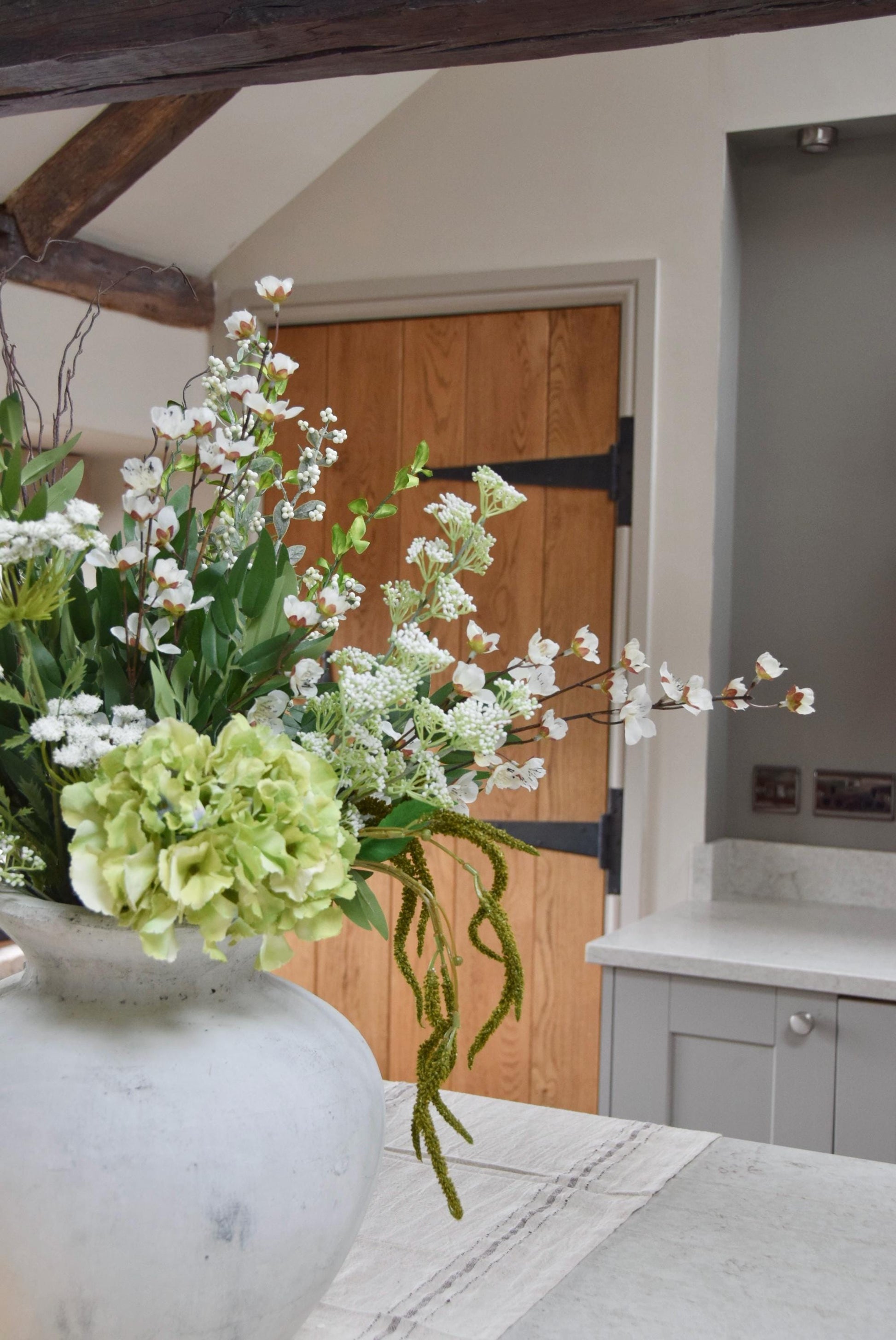 Stone antique vase placed on a kitchen countertop with an arrangement of green and white mixed flowers with lots of foliage. Displayed in a country style kitchen for home decor.