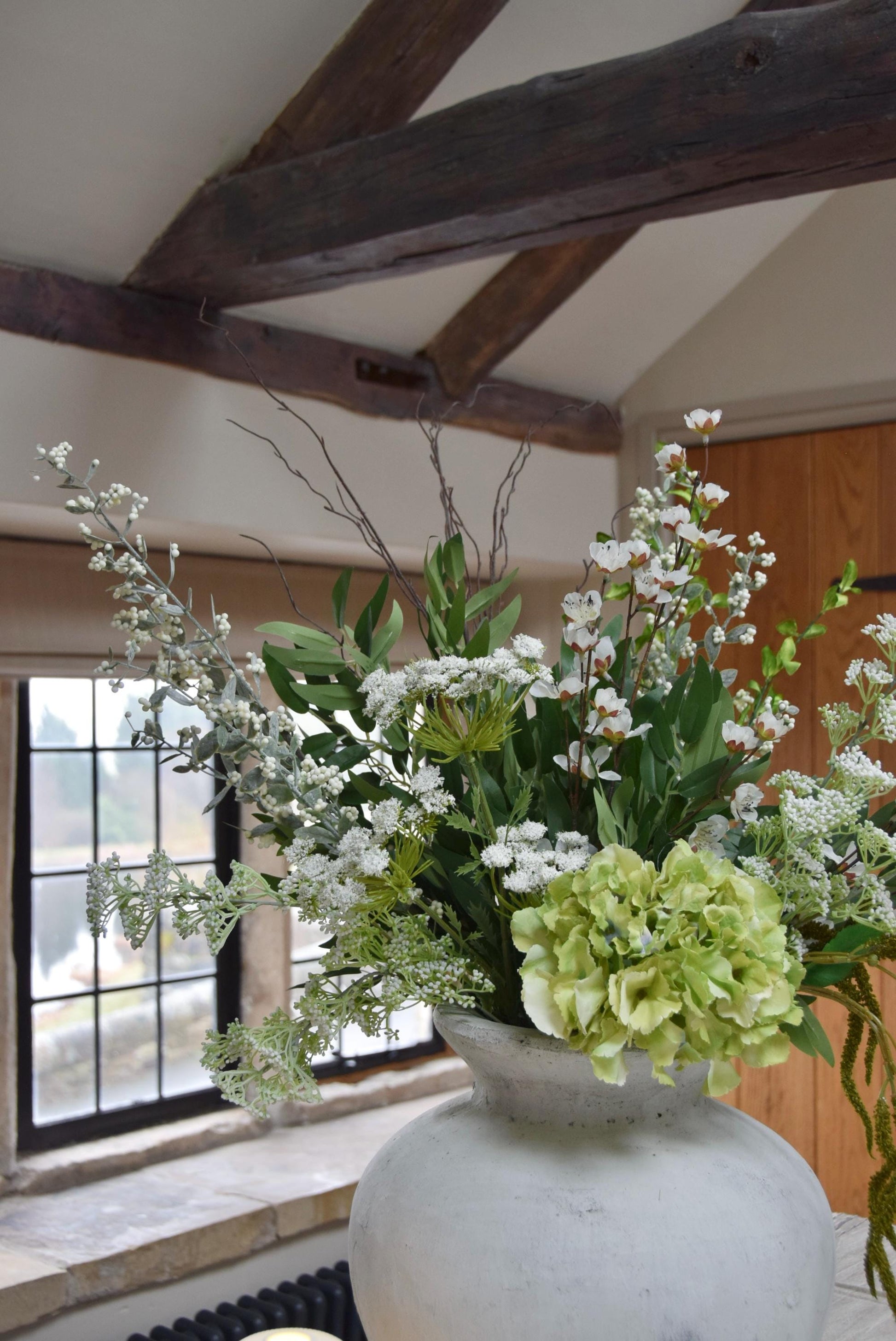 Stone antique vase placed on a kitchen countertop with an arrangement of green and white mixed flowers with lots of foliage. Displayed in a country style kitchen for home decor.