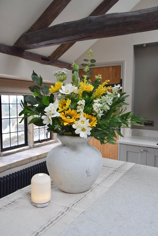 Stone antique vase placed on a kitchen countertop with an arrangement of yellow sunflowers, white wildflowers and green foliage. Displayed in a country style kitchen for home decor.