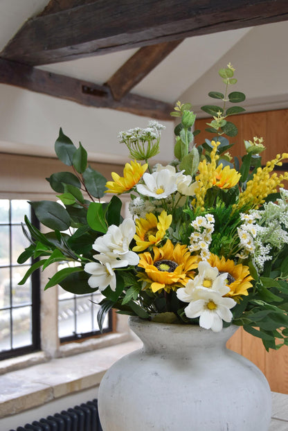 Stone antique vase placed on a kitchen countertop with an arrangement of yellow sunflowers, white wildflowers and green foliage. Displayed in a country style kitchen for home decor.