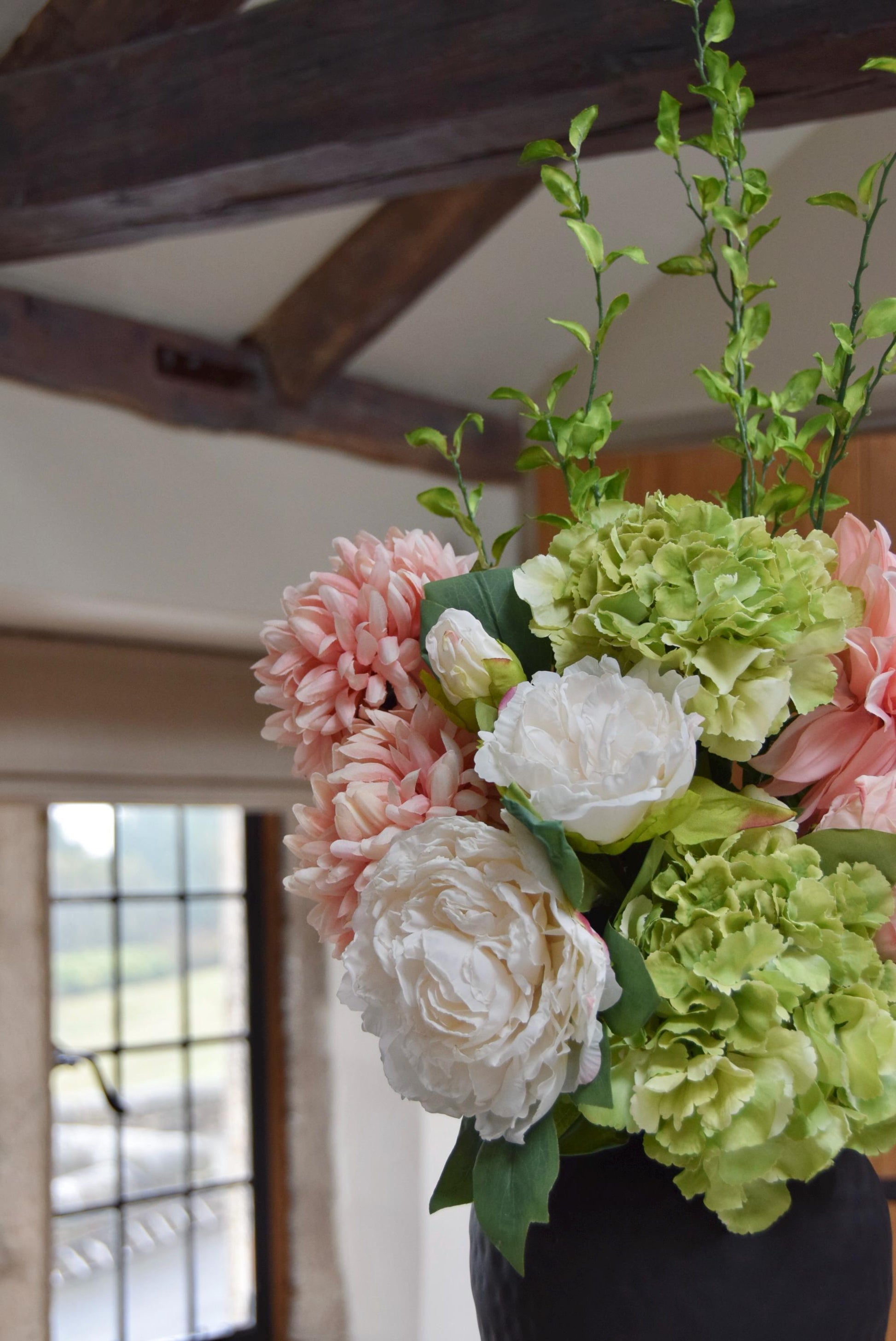 Tall black vase placed on a kitchen countertop with an arrangement of pink and green flowers including green hydrangeas, pink mum flowers, pink dahlias and white peonies with foliage. Displayed in a country style kitchen for home decor.