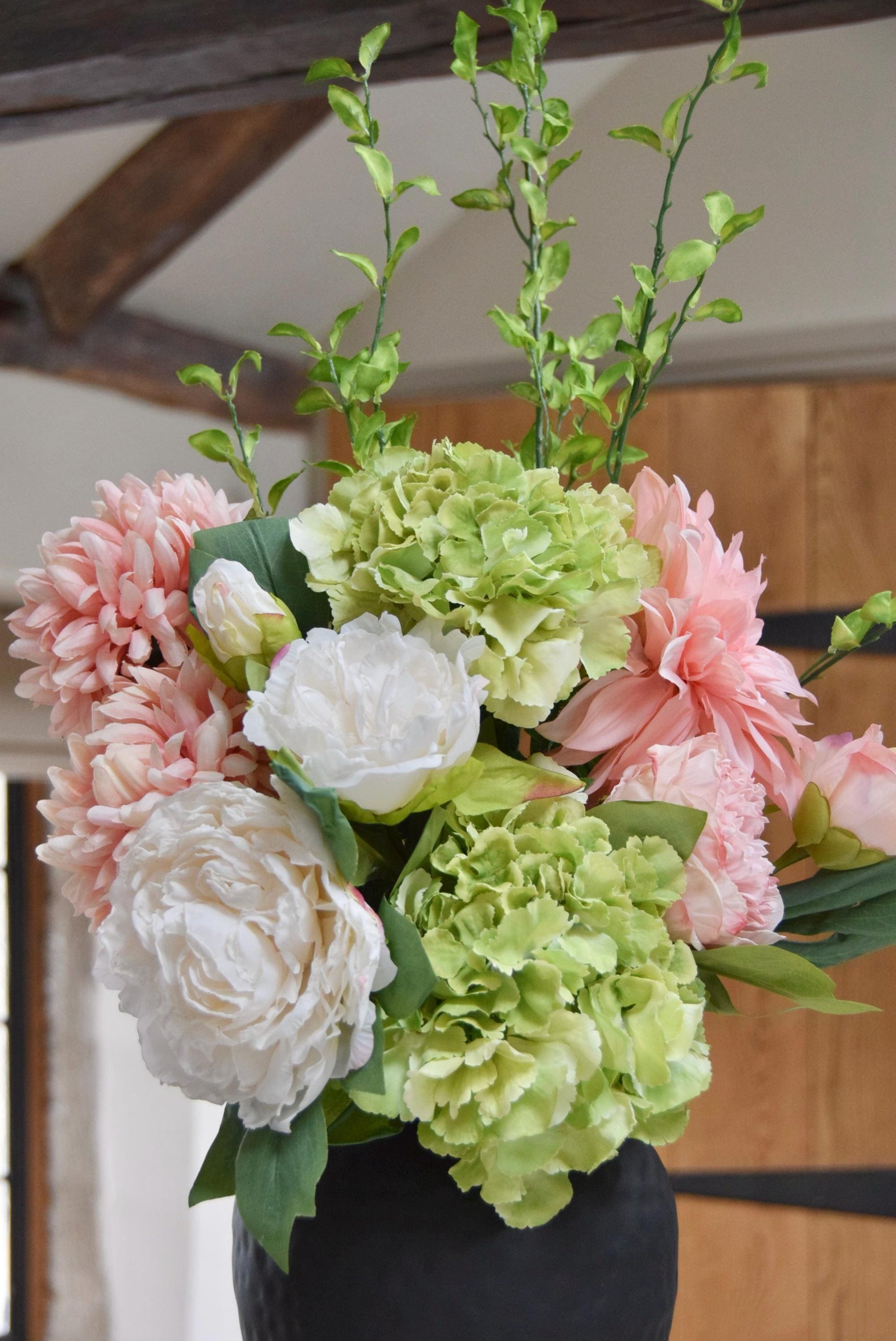 Tall black vase placed on a kitchen countertop with an arrangement of pink and green flowers including green hydrangeas, pink mum flowers, pink dahlias and white peonies with foliage. Displayed in a country style kitchen for home decor.