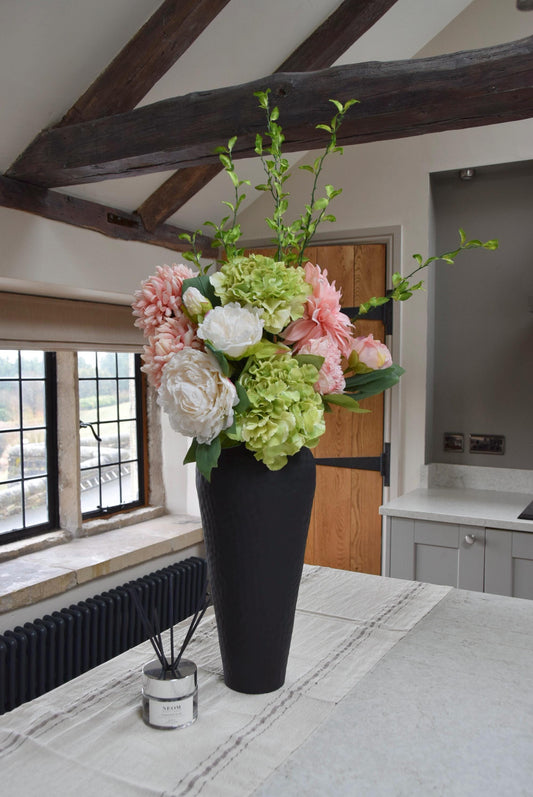 Tall black vase placed on a kitchen countertop with an arrangement of pink and green flowers including green hydrangeas, pink mum flowers, pink dahlias and white peonies with foliage. Displayed in a country style kitchen for home decor.