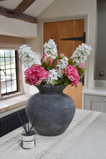 Grey antique vase placed on a kitchen countertop with an arrangement of pink peonies and white paniculata hydrangeas. Displayed in a country style kitchen for home decor.