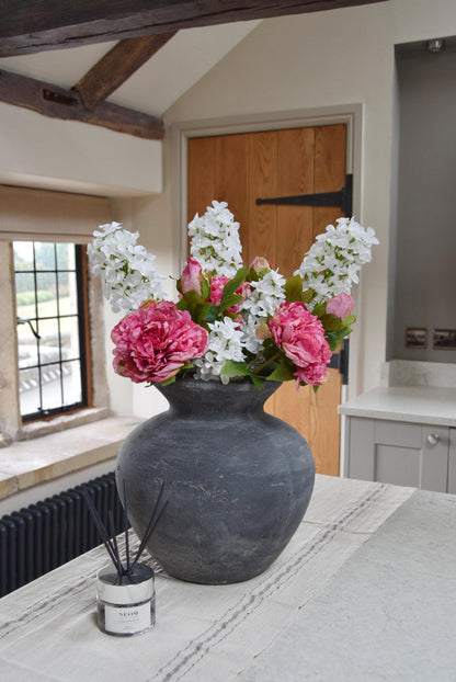 Grey antique vase placed on a kitchen countertop with an arrangement of pink peonies and white paniculata hydrangeas. Displayed in a country style kitchen for home decor.