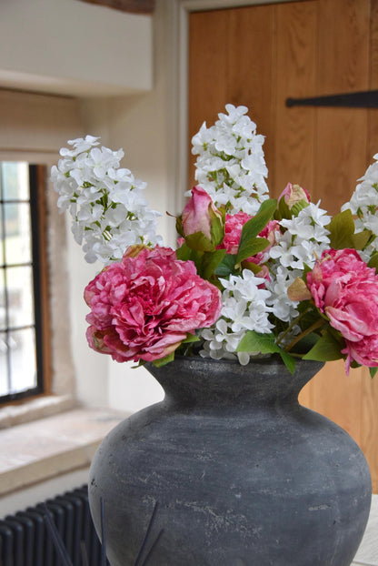 Grey antique vase placed on a kitchen countertop with an arrangement of pink peonies and white paniculata hydrangeas. Displayed in a country style kitchen for home decor.