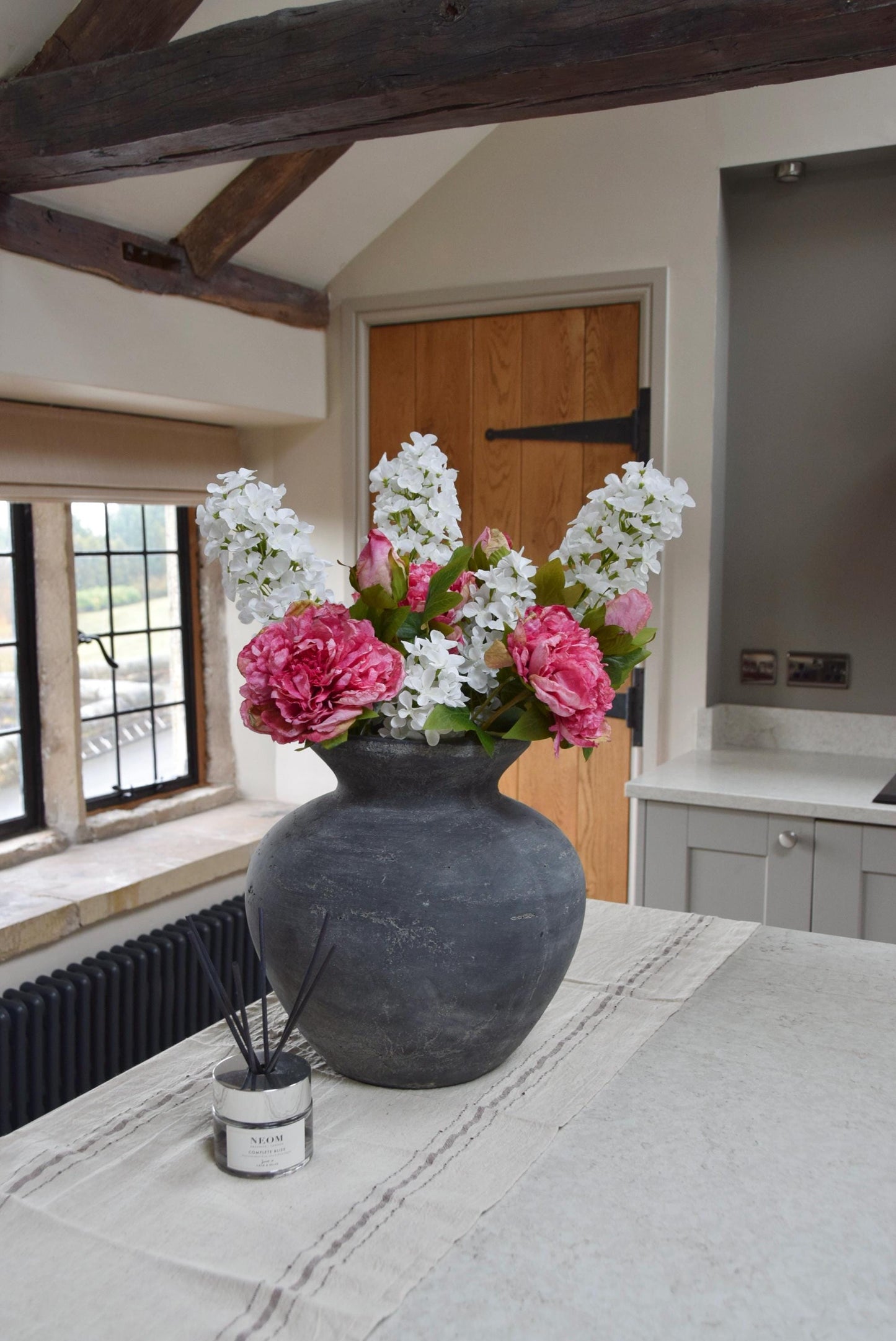 Grey antique vase placed on a kitchen countertop with an arrangement of pink peonies and white paniculata hydrangeas. Displayed in a country style kitchen for home decor.