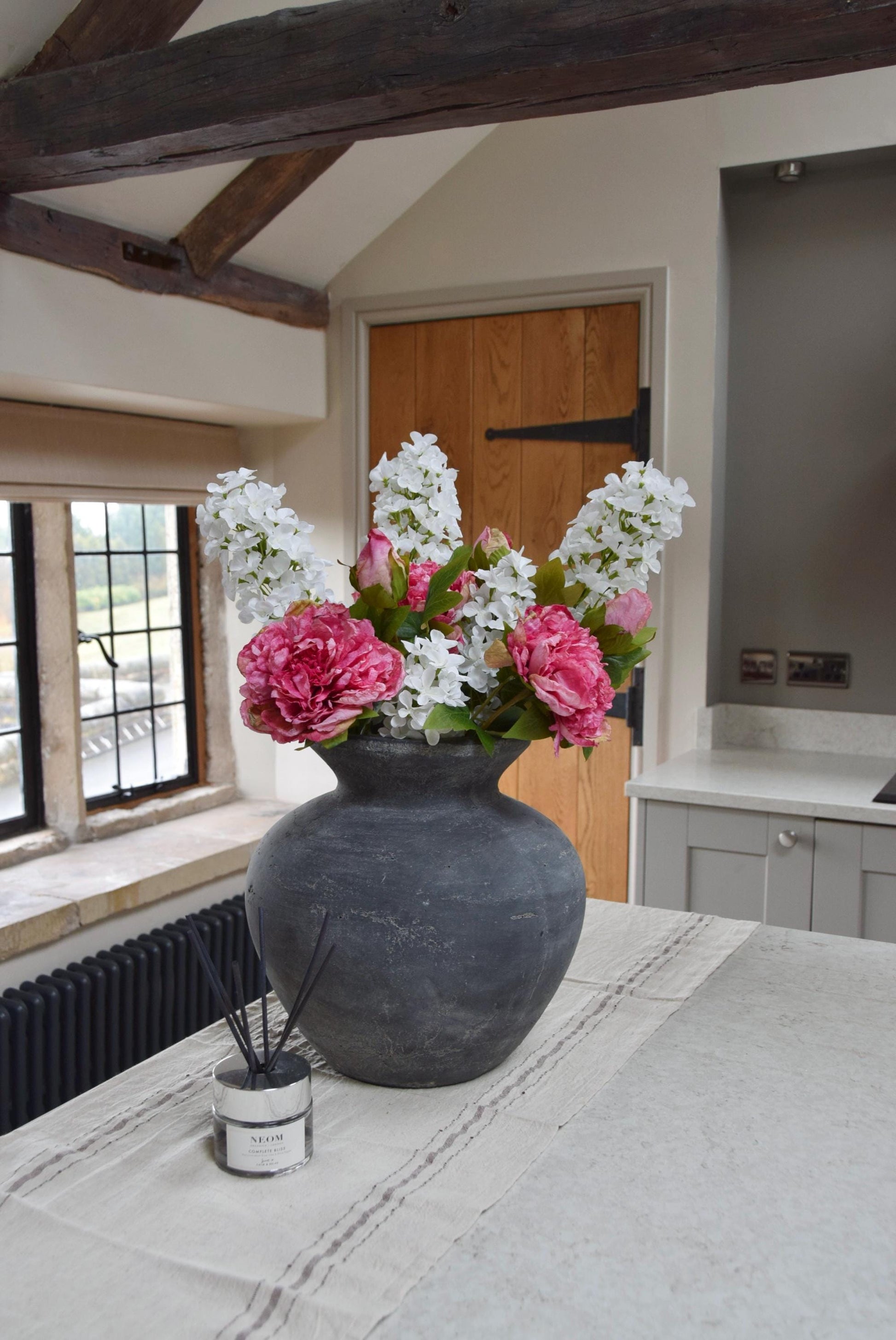 Grey antique vase placed on a kitchen countertop with an arrangement of pink peonies and white paniculata hydrangeas. Displayed in a country style kitchen for home decor.